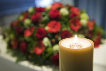 A burning candle with a white coffin and a flower arrangement on the background in a mortuary